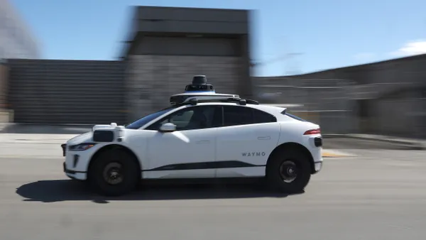 A white vehicles with rooftop and other external sensors, lettered Waymo, drives along a street.