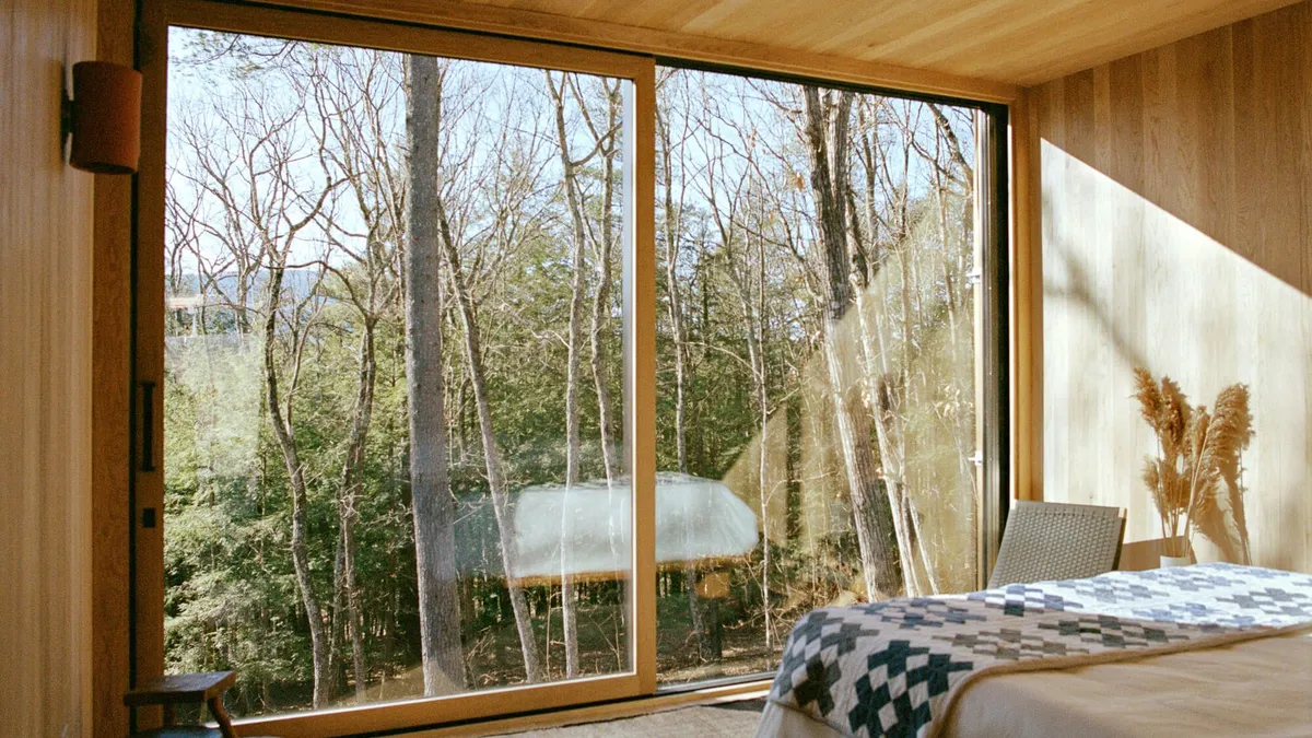 The interior of a cabin features wood-paneled walls and a floor-to-ceiling window overlooking trees.