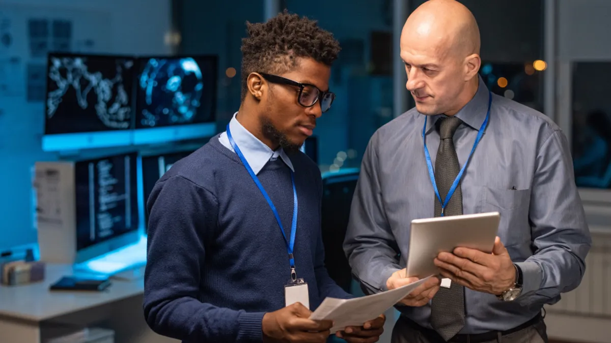 Two elegant intercultural businessmen discussing data in digital tablet held by mature programmer or it-engineer