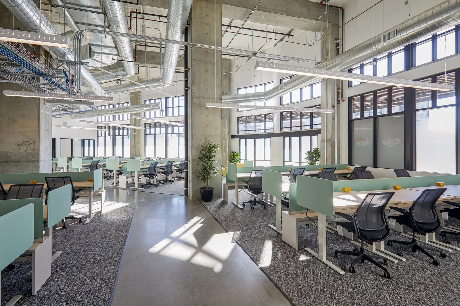 Desk chairs are pushed into grey work tables with light blue sides in a long, light-filled industrial room with ductwork overhead and windows to the right.