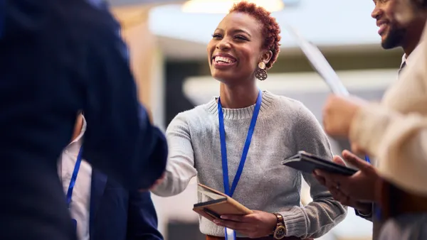 A smiling woman greets another person with a handshake at a professional conference. Other attendees are visible around her.