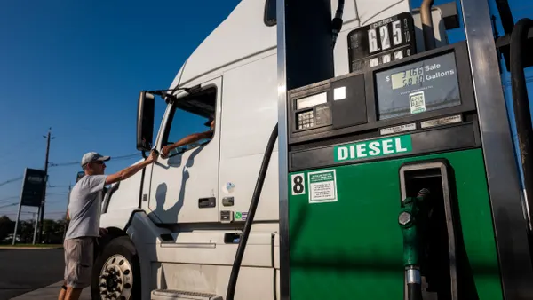 A person on the ground talks to a trucker inside a semi truck while next to a fuel pump showing a diesel price of $6.25 per gallon.