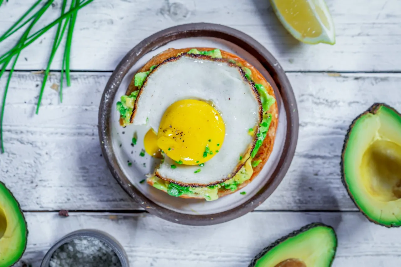 A plant-based fried egg with a liquid yolk on top of an English muffin and avocado, on a table surrounded by avocado halves.