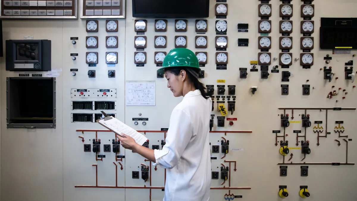 A young woman wearing a blouse and a hard hat working in the control room of a natural gas-fired power plant.