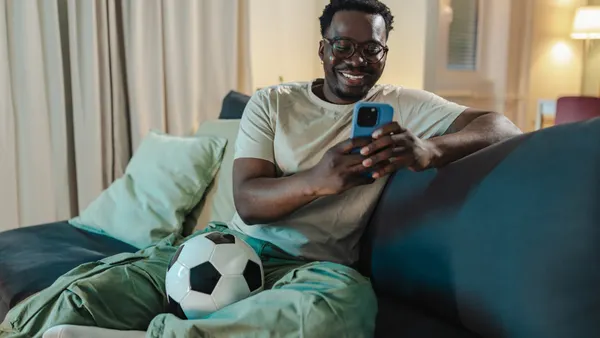 A man with a soccer ball by his feet watches a streaming app on his phone.