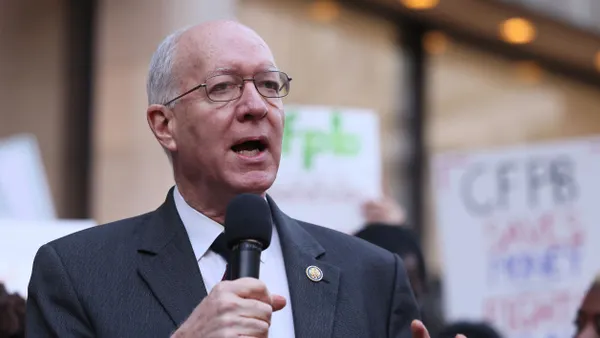 A person wearing a suit and tie holds a microphone while speaking in front of a crowd of people.