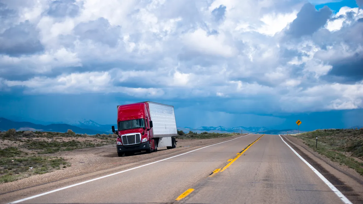 A refrigerated tractor-trailer is stuck on the side of a deserted road with a mountain range in the background.