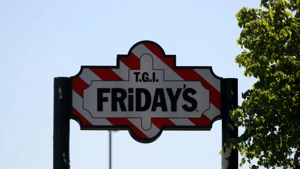 An image ofa sign that says TGI Fridays against oa red and white background. There isa tree on the left and a blue sky in the background