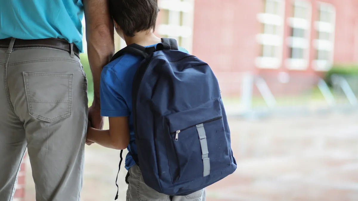 The back of an adult is on the left. The adult is holding the hand of a child carrying a backpack, whose back is also to the camera. The two are standing in front of a building