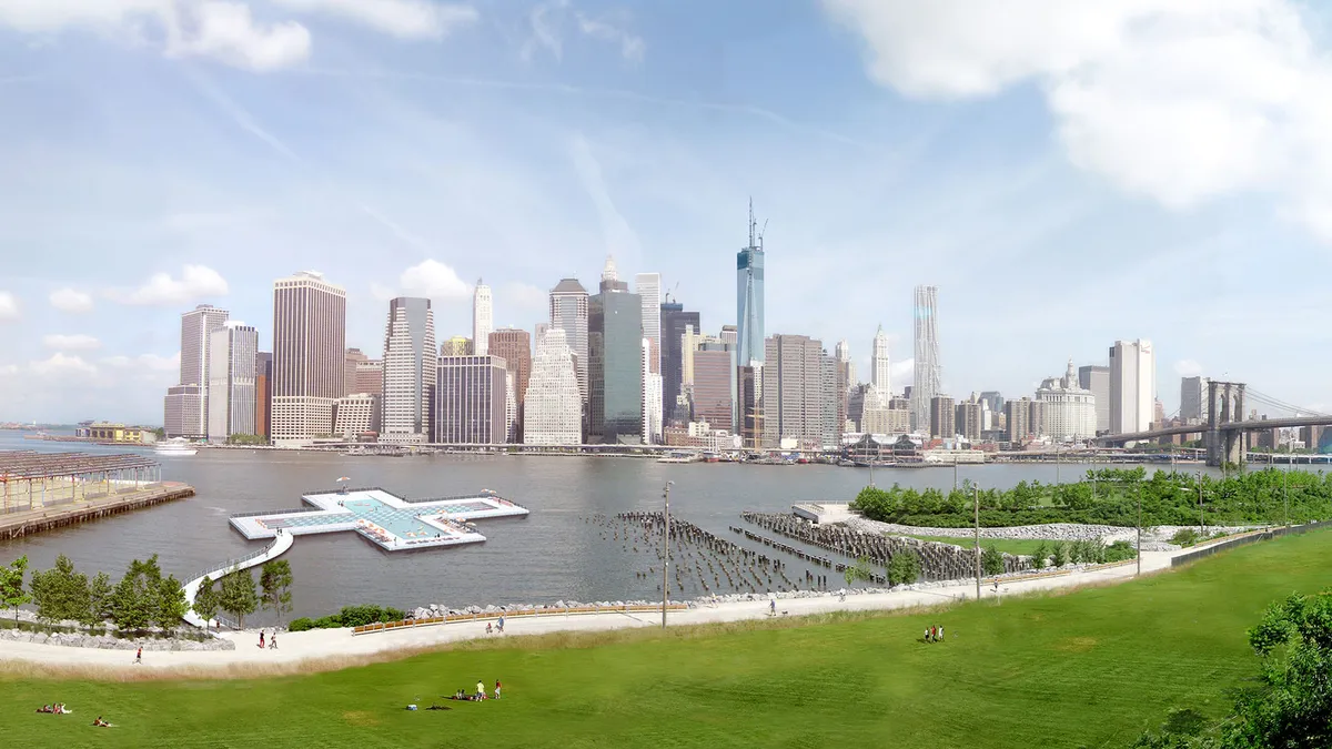 A plus shaped pool in the river in front of a city skyline. A path leads out to the pool from the riverbank.