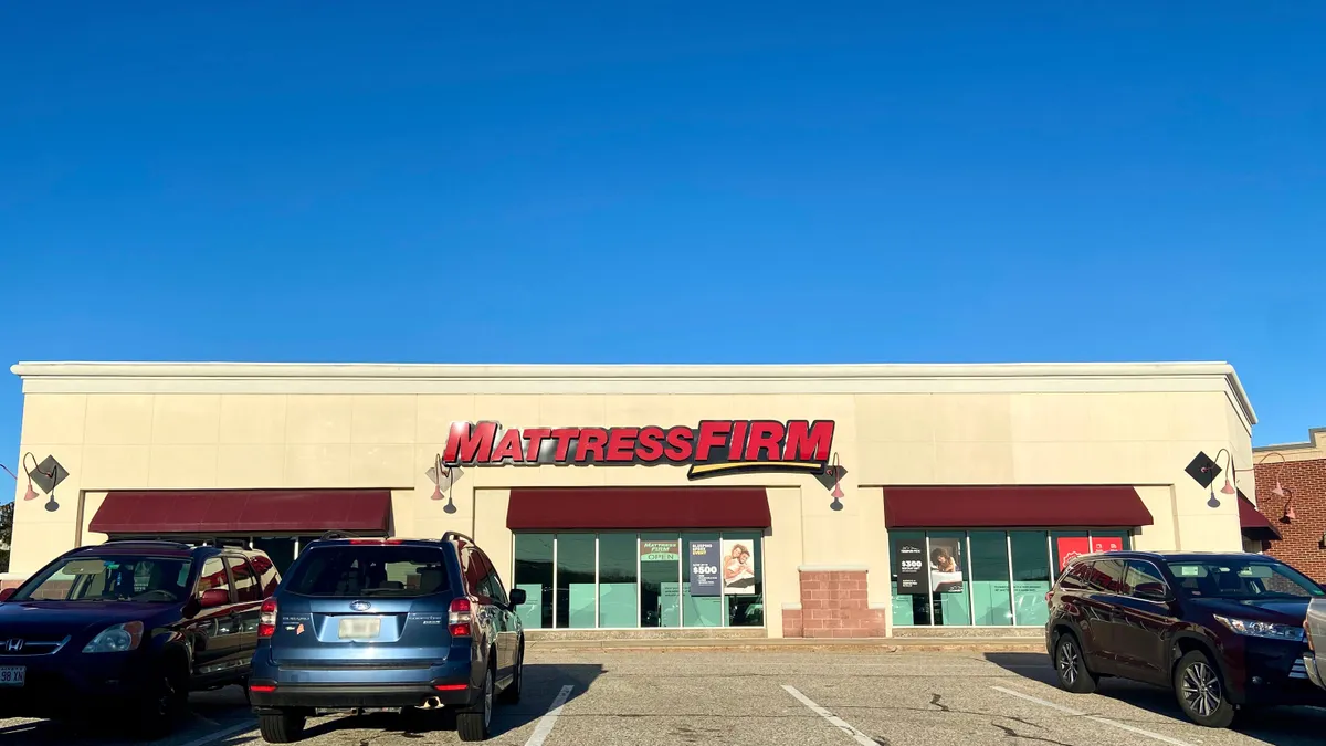 Cars are parked in front of a store with "Mattress Firm" in red capital letters, against a bright blue sky.