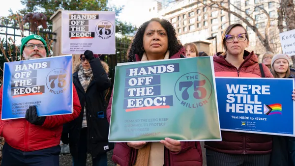 A group of people stand outdoors holding protest signs that read “Hands Off the EEOC” and “We’re Still Here,” with one sign featuring a rainbow-colored arrow, as buildings and trees appear in the background."