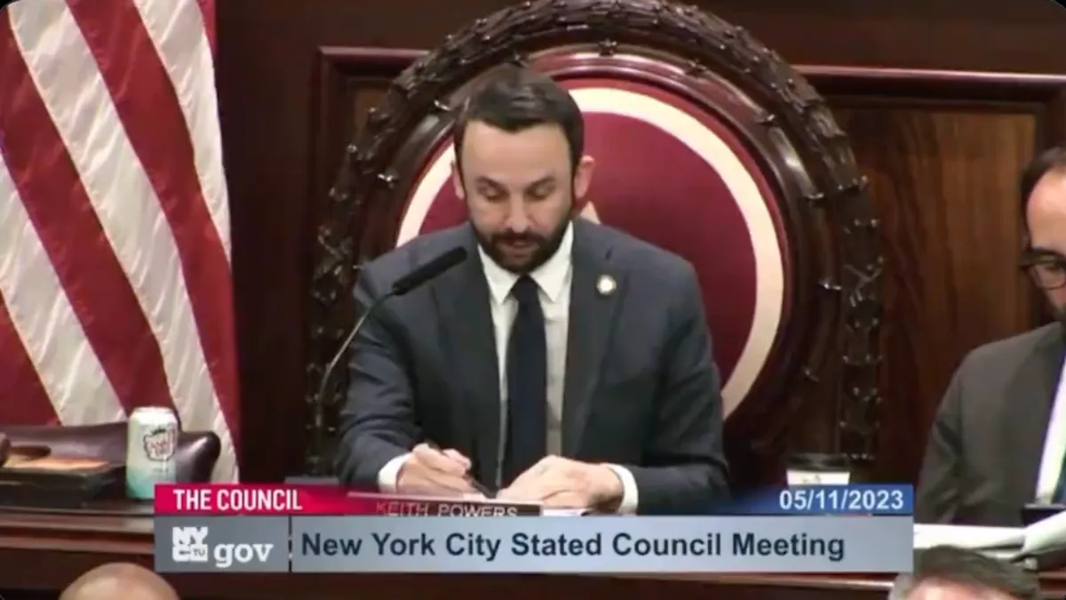 Keith Powers, of the New York City Council, presides over a meeting with an American flag to his left and a man seated to his right.