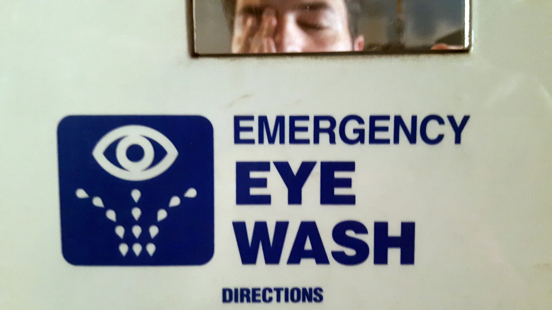 A blue and dark white emergency eyewash sign, with a small rectangle mirror reflecting a pereson washing their eyes.