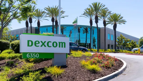 Modern office building with tall glass windows and palm trees lining the entry drive. A large white and green sign in the foreground reads “Dexcom” with the number “6350” below. Surrounding the sign are colorful landscaping features including small bushes, flowers, and a mulched bed. Cars and flags are visible in the background.