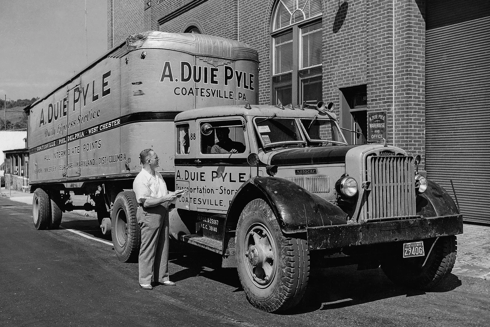 A historic photo of a person speaking to another person sitting in a large supply truck. The branding on the truck door reads, “A. Duie Pyle, Transportation - Storage, Coatesville, PA”.