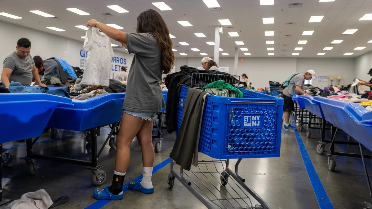 A person finds a T-shirt from a Goodwill outlet bin and holds it up to examine it. Next to the person is a cart filled with other clothing items.