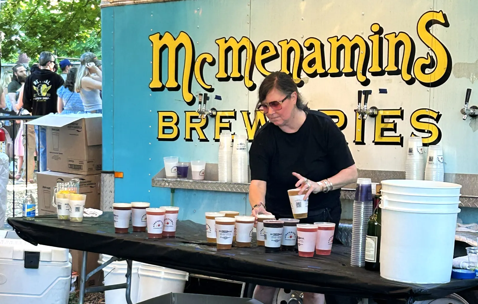 A person stands behind a table with beverages in cups on it, and a McMenamins Breweries sign behind them.
