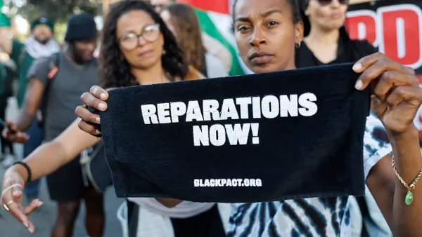 A protester displays a sign reading "Reparations Now!" in front of a crowd of other protesters.