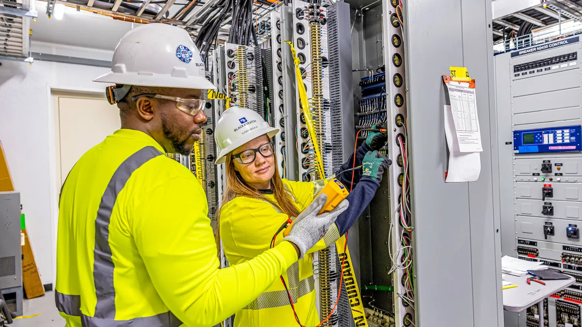 Two electrical workers in high-visibility gear work on wiring inside an electrical panel in a control room