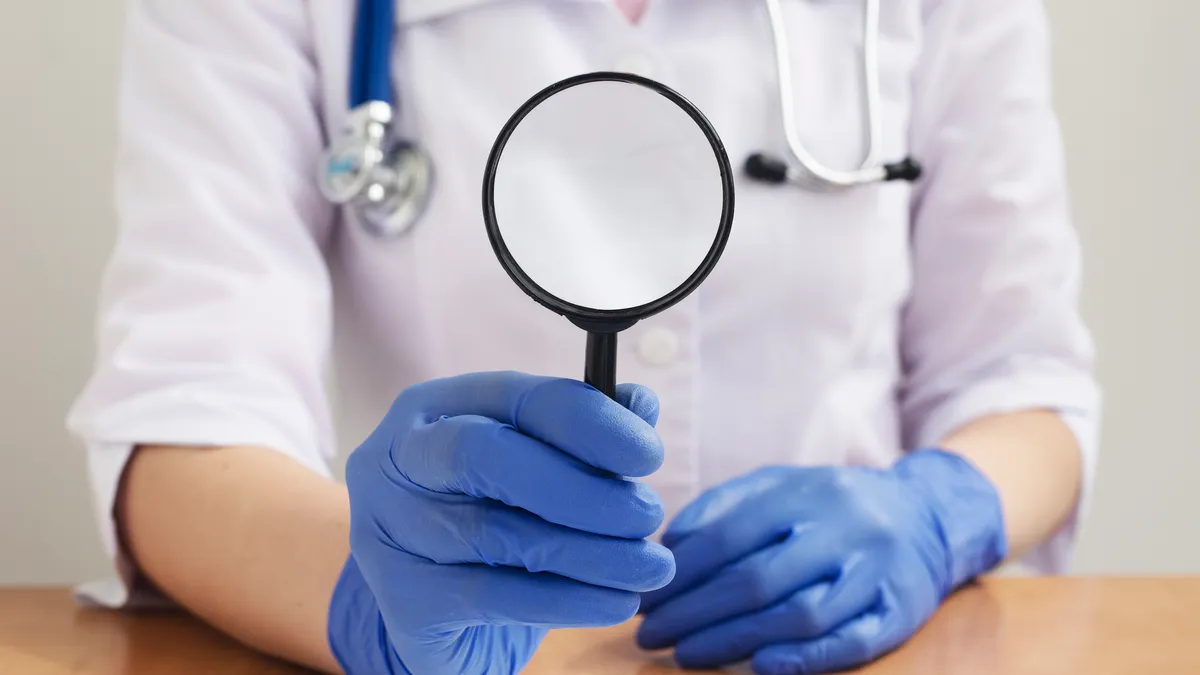 A person in a white doctors coat and stethoscope holds up a magnifying glass.