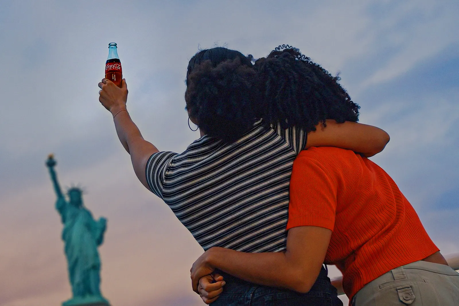 Two people embrace in front of the Statue of Liberty while one holds a Coke