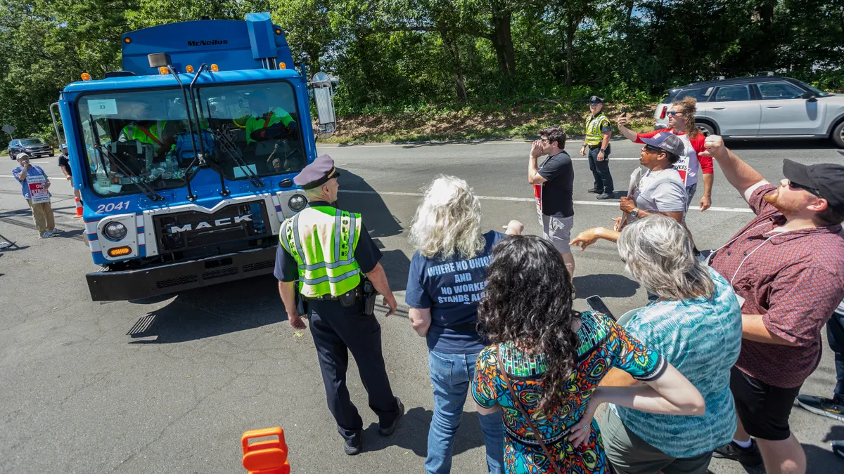 Several people holding or wearing signs stand shouting in front of a garbage truck, watched by two police officers in high-visibility vests.