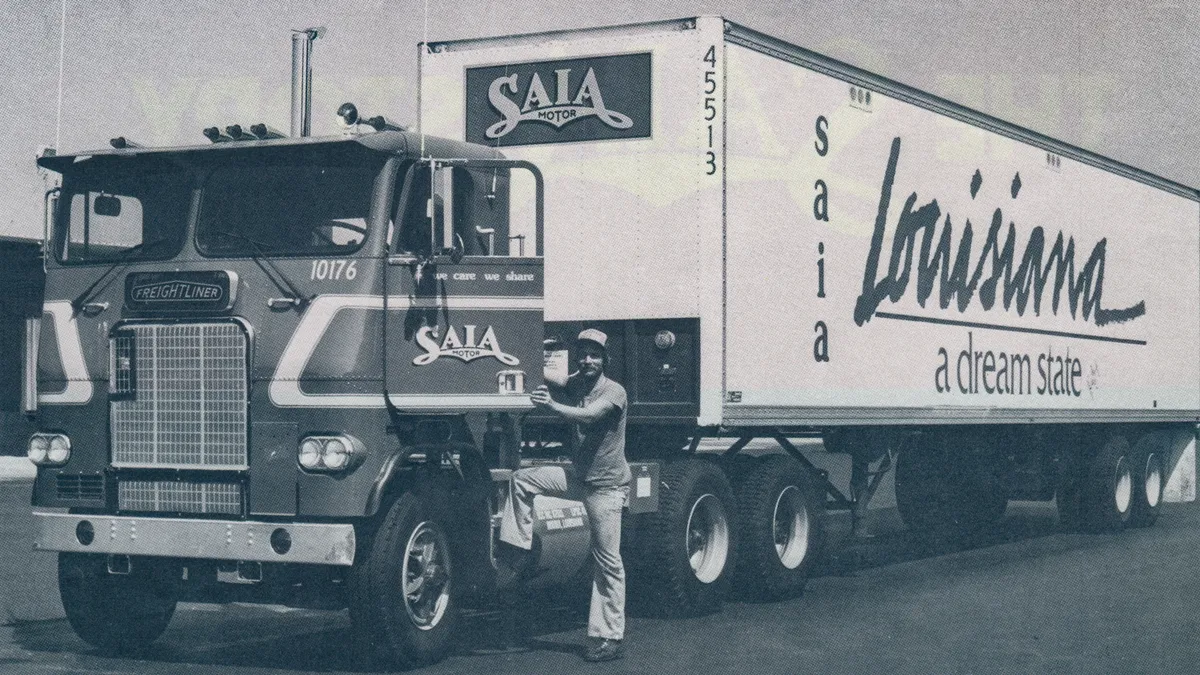 A person stands with their foot on the first step of a Saia Freightliner truck attached to a white trailer labeled “Saia, Louisiana, a dream state” in a black-and-white photo.