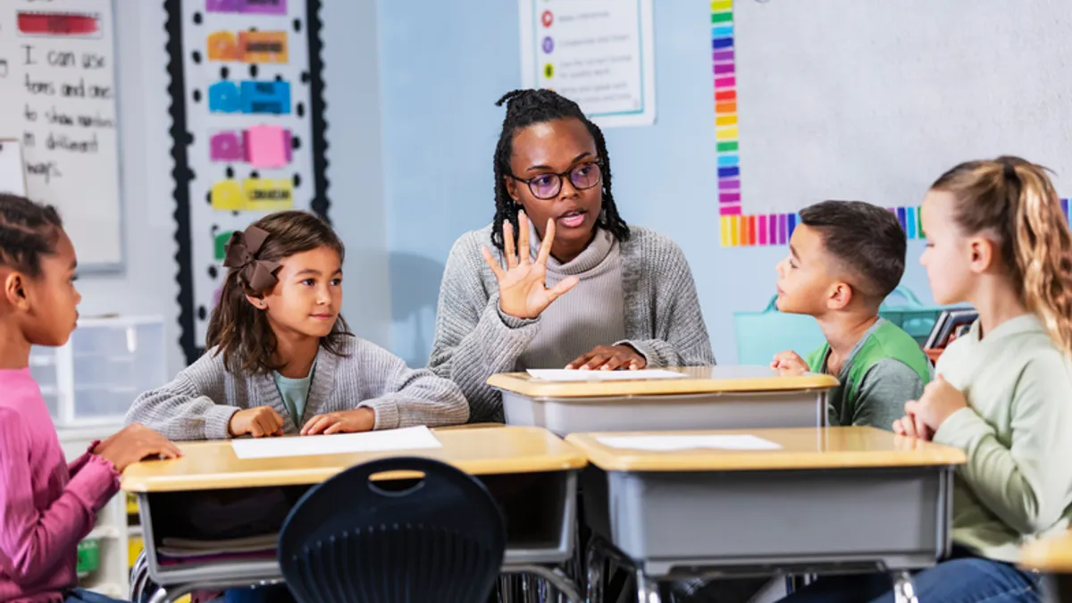 An adult is sitting at a desk in a classroom with four young students who are sitting at desks close together. The students are looking at the adult who has their hand up and five fingers out.