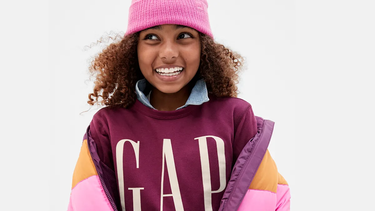 A smiling tween is wearing a pink hat and maroon sweatshirt that says "Gap," underneath a pink and gold winter ski coat.