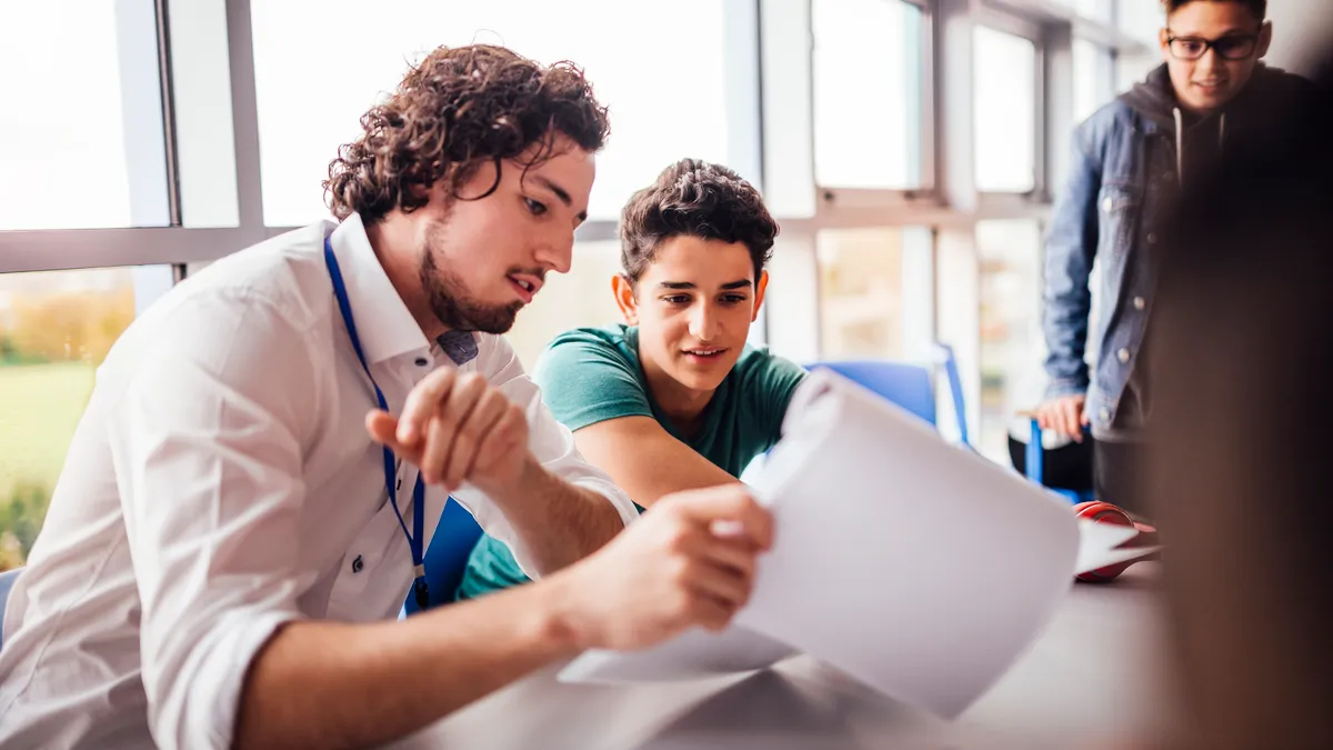 An adult sits at a desk next to a student. The adult is holding papers and both are looking at papers. Another student looks on.