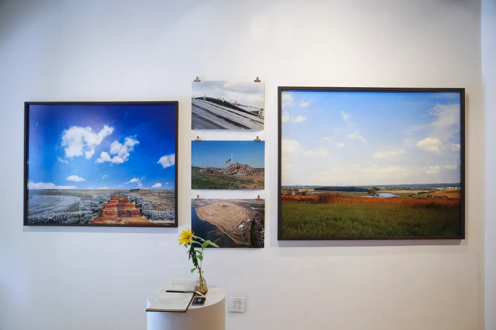 Two large prints, one featuring a manmade water flow and the other showing a natural landscape with the New York City skyline in the distance, hang on a gallery wall.