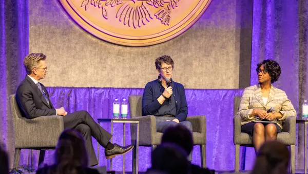 Three people are sitting in chairs on a stage. One person has a microphone