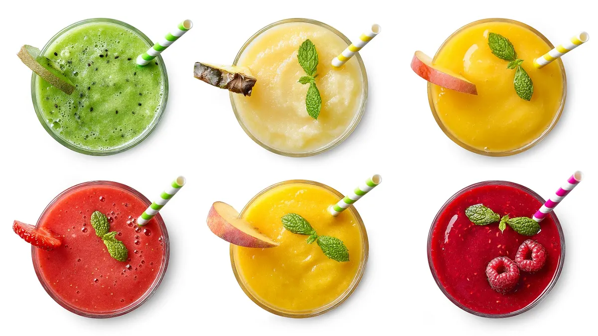 Overhead view of six glasses of fruit juice garnished with fruit and mint and with a straw. Clockwise from top left: kiwi, pineapple, apple, raspberry, peach, strawberry