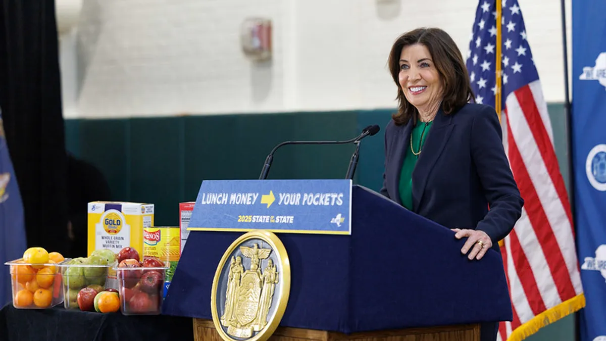 New York Gov. Kathy Hochul stands behind a podium next to a variety of food items as she announces a universal school meals proposal in a school cafeteria.