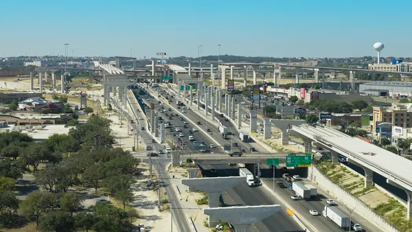 Highway interchange in Texas with concrete support columns and partially completed elevated ramps rising above active traffic lanes, with semi-trucks and other cars navigating lanes.