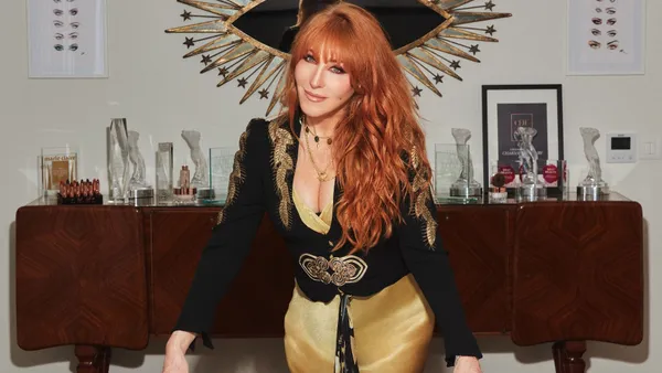 A woman with long red hair leans on a table covered in beauty products and faces the camera.