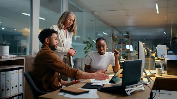 Two people are sitting while one is standing, reviewing something on a laptop.