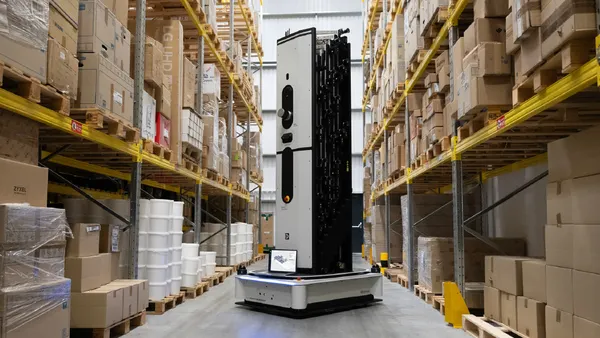 A tall autonomous robot navigates a warehouse aisle lined with shelves of boxes and goods.
