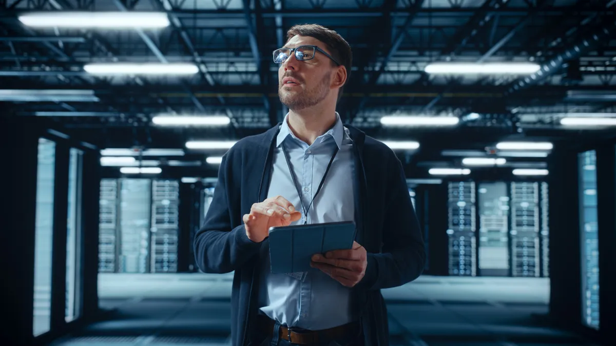 Male IT Specialist Walks Between Row of Operational Server Racks in Data Center.