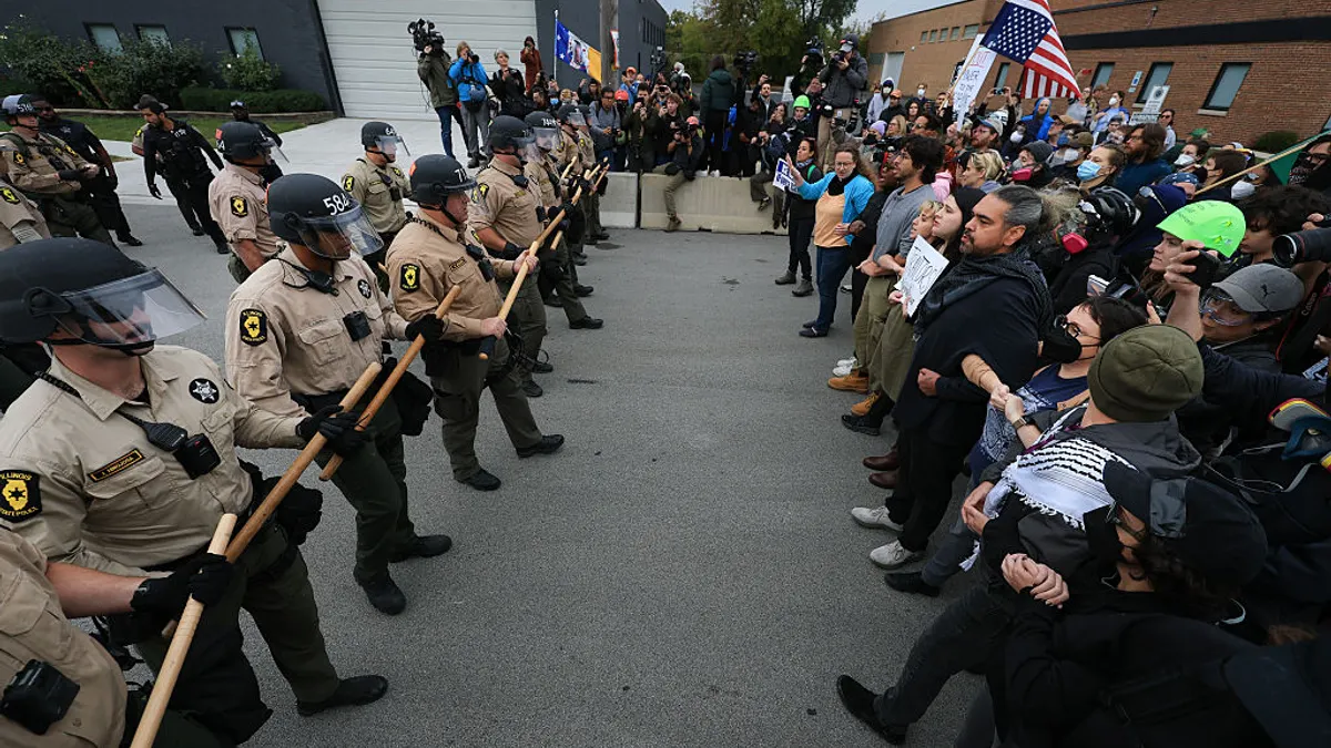 two groups of people are standing on a street facing each other. One side has posters and the other side is dressed in law enforcement uniforms.