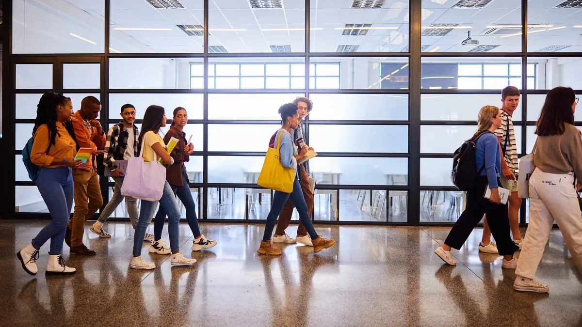 A group of students are walking in a hallway with a large floor to ceiling window. Their sides are shown and they are carryign books and wearing backpacks.