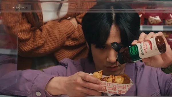A person is holding a small container of food and squeezing sauce onto the food. Another person stands behind him with their arms crossed. There is a  set of shelves with bags of packaged goods on it behind them.