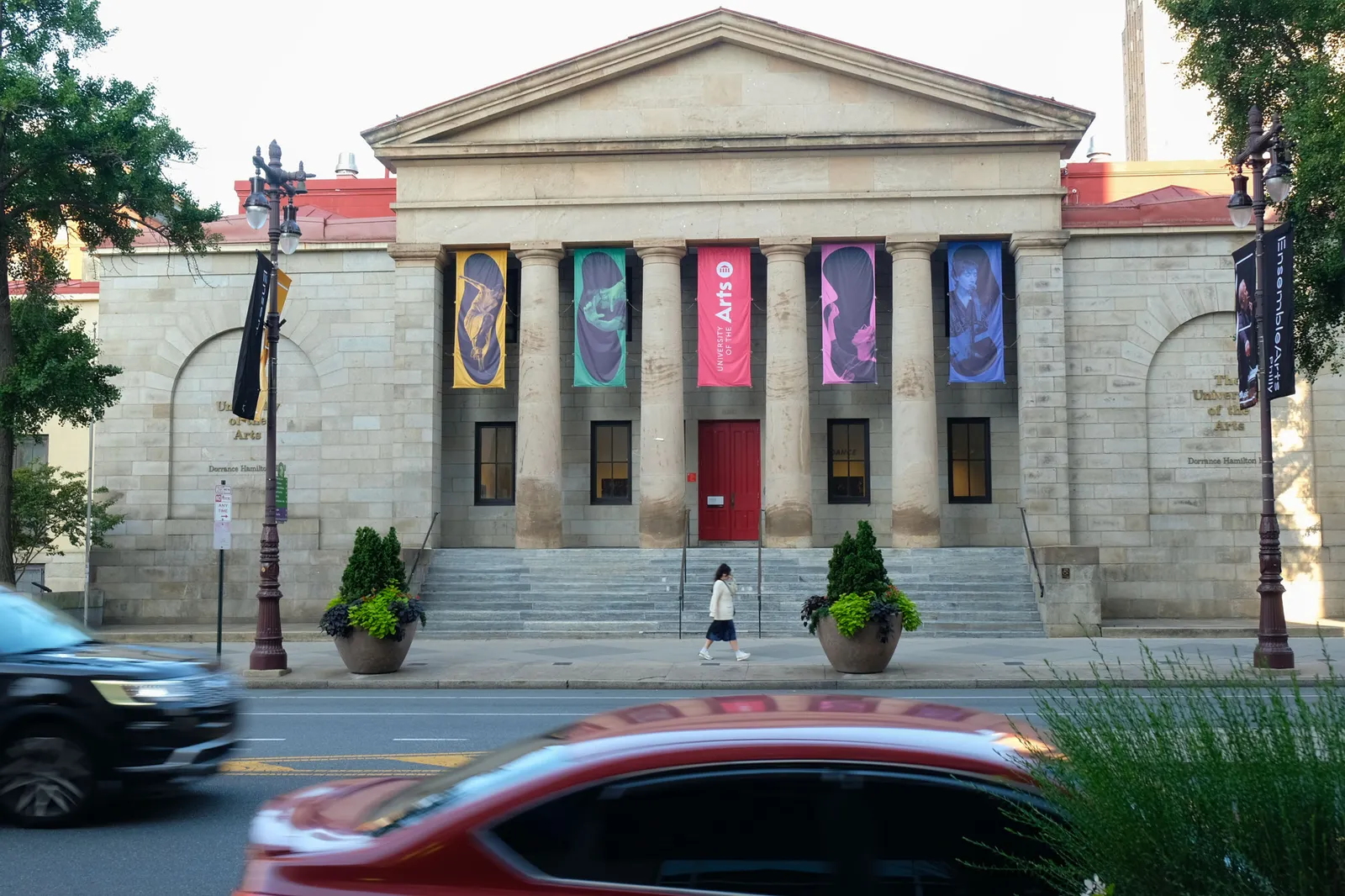 View of building with columns and colored banners as vehicles and a pedestrian pass.