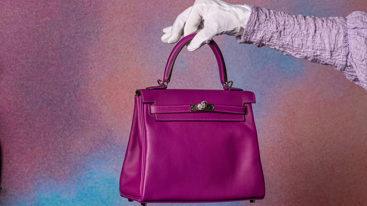 A white-gloved hand holds out a purple Hermes Kelly bag.