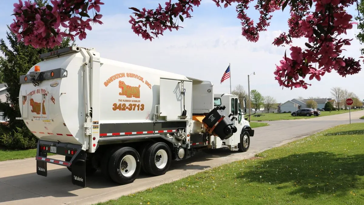 A garbage truck with Sanitation Service Inc., branding and a logo drives down a street in front of a blossoming tree.