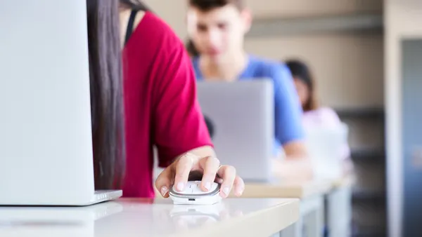 Students siting in rows of desks are using laptops and a mouse. Only the sides bodies of two students are shown in the photo.