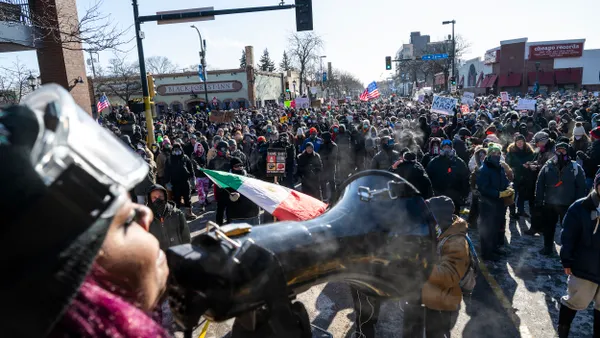 A person wearing a helmet speaks into a bullhorn to a large crowd holding signs.