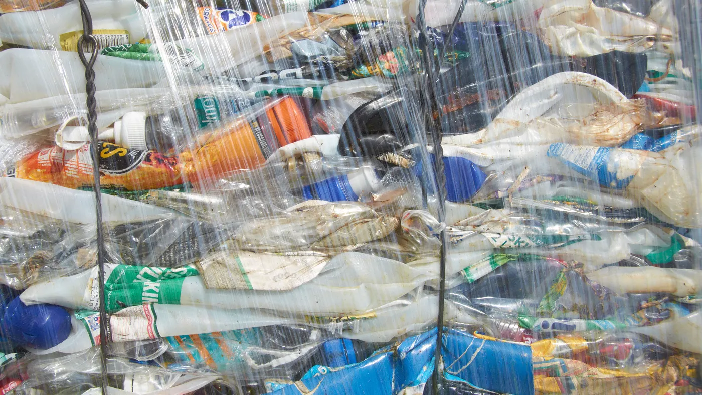 A tightly packed bale of crushed plastic bottles, compressed into a rectangular block and wrapped in clear plastic film. The bale contains various colored plastic bottles, flattened and compacted together, ready for recycling.