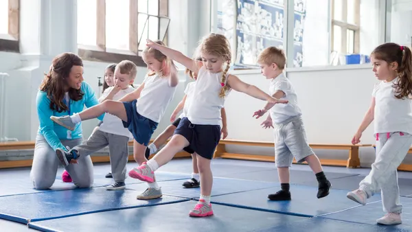 Young students are in a large room and are trying to balance on one foot while standing on mats. An adult is helping one student.